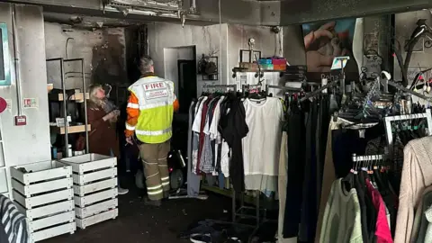 Whitby Wildlife Sanctuary The interior of a shop that appears to have recently suffered fire damage.
The ceiling, walls, and parts of the back room are blackened with soot and smoke stains. A firefighter wearing a high‑visibility jacket marked “FIRE” is standing inside the shop. Clothing racks filled with shirts, trousers, and sweaters are still standing in the foreground, contrasting with the burned area toward the back. 