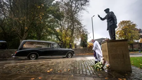 PA Media The funeral cortege for Dickie Bird passes the life-size bronze statue of Dickie Bird near St Mary’s Church, Barnsley.