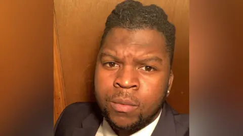 A selfie of a man with dark hair and a short dark beard, wearing a black and white suit.