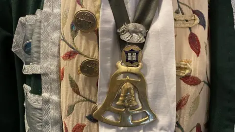 A close-up of Mr Ward's regalia and town crier medal. His robes are dark green and silver. He has a beige shirt with colourful leaves on them. The medal is on a dark green ribbon and is gold and in the shape of a bell. There is a crest above the bell and a silver pig brooch above that. 