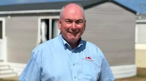 Patrick has grey hair and wears a light blue shirt - he stands with a static caravan behind him.