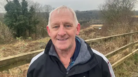 John Ashworth, smiling wearing a tracksuit, stands in front of the derelict Nob End locks on the Manchester, Bolton and Bury Canal in the background.