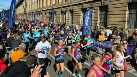 A sea of runners jog down a main street in Hull wearing colourful clothing while crowds of spectators cheer them on. There are 'Run for All' flags lining the pavement. It look to be a warm, sunny day.