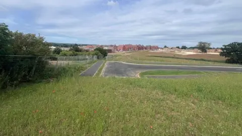 A field with an unfinished road running through and across it. In the distance is a housing estate