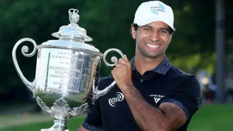 Rai holds a massive trophy and is smiling at the camera. He is wearing a white cap and navy top.