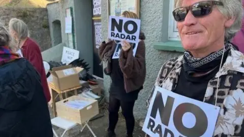 A group of protesters holding signs saying "no radar", stood next to a house.