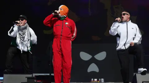 The band Kneecap perform onstage - all three members of the band are holding microphones. One of the group, standing centre, is wearing all red with a small Irish flag printed on it and a tricolour balaclava. The other two band members are wearing white jackets and sunglasses.