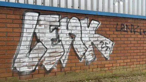 White graffiti on a light brown brick wall, which says 'lenk'. To the right of it is a small patch of graffiti written in black ink, which says 'lenk'