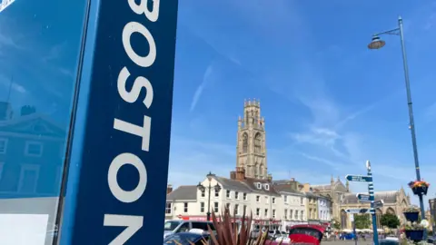 A view of a town market place, dominated by a large medieval church with a tall tower under a blue sky. In the foreground, a blue vertical sign on the side of a bus shelter reads "Boston" in white letters.