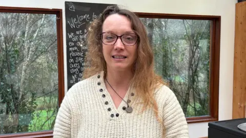 Linzi has long light brown hair and glasses and wears a cream coloured knitted top while standing in front of some windows and a blackboard in the cafe 