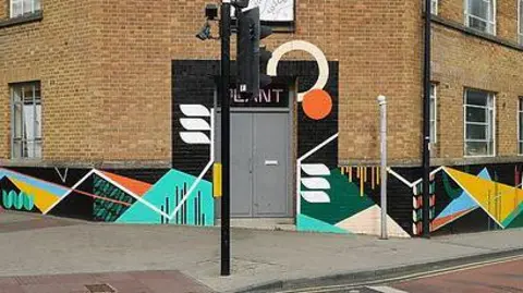 Supplied A geometric mural with black, turquoise, white, orange, and yellow shapes decorates the lower exterior walls of a brick corner building. The mural wraps around the street-facing corner beside a grey doorway and is bordered by a pavement, traffic lights, and a bicycle lane.