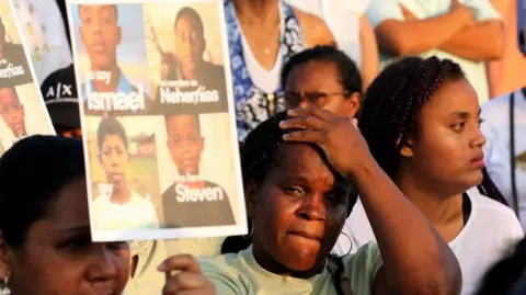 JONATHAN MIRANDA/EPA/Shutterstock A person reacts during a tribute dedicated to Ismael and Josue Arroyo, Nehemias Arboleda and Steven Medina in Guayaquil on 8 December 2025. The woman clutches her forehead while holding up a composite picture of the four boys. Around her are other women also taking part in the tribute. 