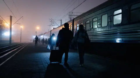 BBC/Matthew Goddard A train station in the mist. You can see a cluster of people on the station platform. 