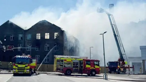 Three fire engines are outside a burning building, which looks like an old mill and is missing a roof 