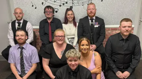 Supplied A family photo showing a mixture of nine men and women wearing either smart shirts and ties or dresses. They are positioned close together in front of a wall and they are all smiling while looking into the camera. 