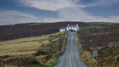 MANX SCENES The road leading from Creg ny Baa with a grass embankment on the right, and green hills on the left. a white cottage is straight ahead on the left side of the road.