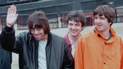 PA Media Oasis offstage at their Knebworth shows in 1996 - Liam in tinted glasses raising a palm, with Noel Gallagher next to him and Paul McGuigan behind them