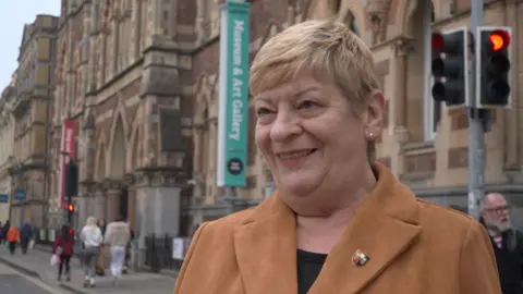 Laura Wright, who has short blonde hair and is wearing a tan coloured coat, stands in front of the RAMM Museum and Art Gallery in Exeter. The building is red brick, with arched windows and columns.