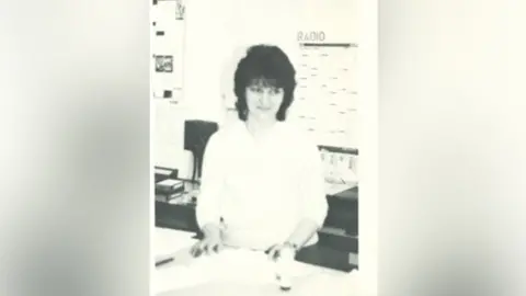 Sharnbrook Upper School Pat pictured in black and white standing with her hands on a photocopier or desk and glancing slightly to one side. She wears a white blouse and has black curly hair in a bob.