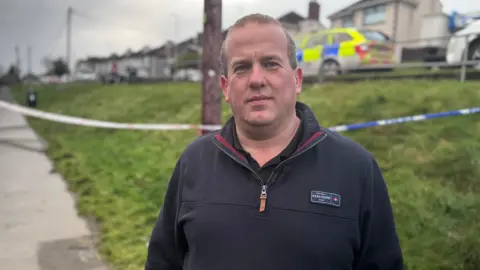 Clement Cuthebertson has short fair hair, and is wearing a quarter zip pullover. Behind him is a grass bank, and beyond, a police car parked on the road. 