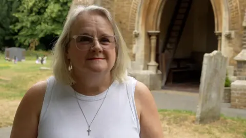 John Fairhall/BBC A woman wearing a white vest top with a crucifix necklace on. She has blonde hair and is standing at the entrance of a church.