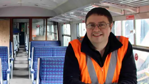 A smiling Dan Bennet inside the old railway carriage with its blue and purple patterned seats. He has short dark hair, is wearing black rimmed glasses and is wearing an orange high-viz vest.