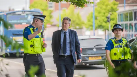 West Midlands Police and Crime Commissioner A man in a navy blue suit with a light blue shirt and a navy blue tie is walking whilst talking with two police officers. They are on a pavement with a bus and a cars on the road beside them. There are trees and buildings in the distance.