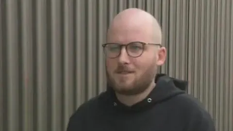 RTÉ Stephane Benz - a bald man with a beard is wearing circular glasses as he walks in front of a green corrugated steel fence.