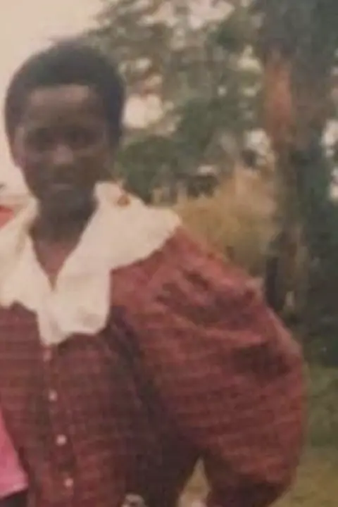 Emertha Uwanyingira Emertha aged 12. She has short hair and is wearing a burgundy shirt dress with a white check pattern and white lace collar and sleeves. There are grass and trees in the background and she is smiling at the camera.