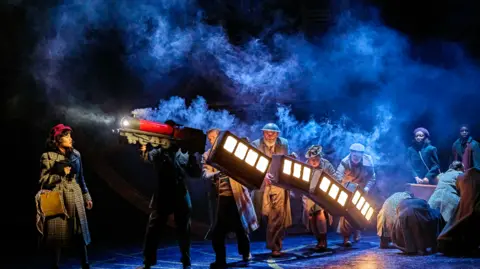 Brinkhoff-Moegenburg Actors carry an illuminated model train across a dark, smoky stage