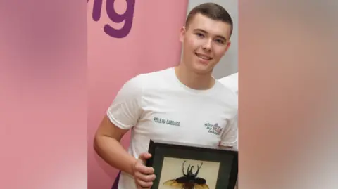 Glór na Móna A young Liam Óg Ó hAnnaidh, known by his stage name Mo Chara, smiles at the camera. He is wearing a white tshirt with the Glór na Móna logo on one side and Féile Na Carraige on the other. He is holding a framed bug and is standing in front of a pink backdrop.