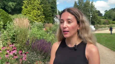 Piers Hopkirk/BBC A blonde woman, with a small microphone attached to her black top, is standing next to some flowers and plants at a park. 