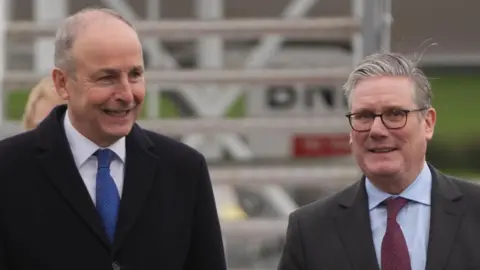 Micheal Martin, a man with grey hair, a black coat, and blue tie and Sir Keir Starmer, a man with grey hair, glasses, grey suit and claret tie at the airport in Cork.