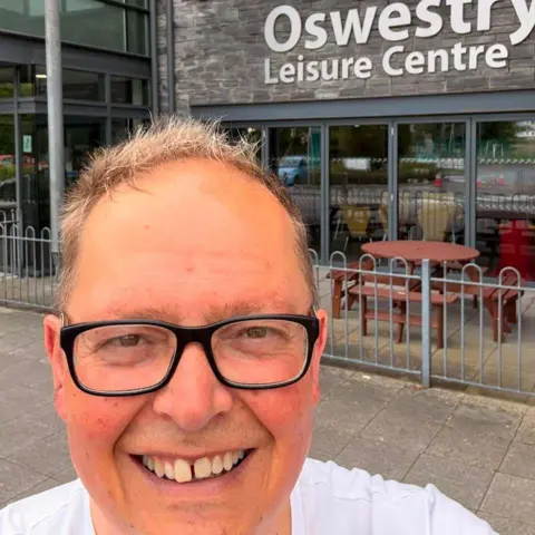DAN WILLIAMS Dan Williams, who has short brown hair and is wearing glasses, with Oswestry Leisure Centre behind him.