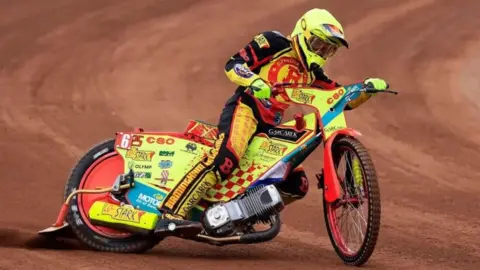 Getty Images A person riding a speedway motorbike on a brown dirt track. The bike is yellow with red paint and dozens of sponsor logos over the body of it. The rider is wearing a red, black and yellow race suit with a yellow helmet and gloves