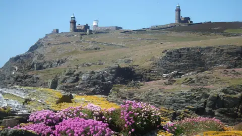 ARON SAPSFORD A number of buildings on a hill. There is grass and steep rock faces with pink flowers in the foreground.