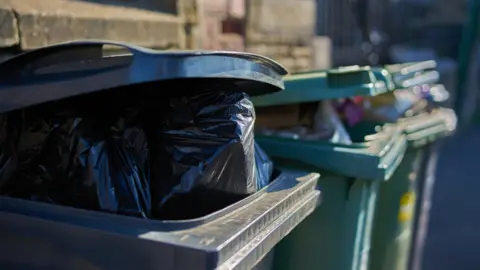 Some wheelie bins filled with black bin bags, forcing the lids open.