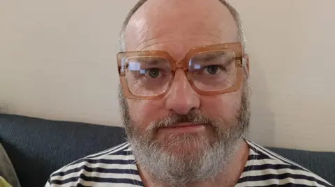BBC A man with short grey and a grey beard is wearing large brown square glasses. He is sitting on a grey sofa in front of a cream wall and wearing a black striped top.