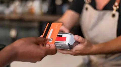 Getty Images Contactless card payment being made with a card held in a hand and a terminal being held by someone in an apron.