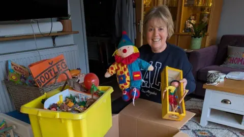 Angie Emery A woman with short blonde hair wearing a dark blue jumper holding up children's toys, she is surrounded by boxes of children's toys. 
