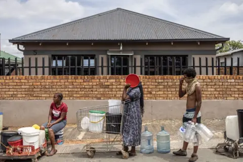 Roberta Ciuccio/AFP Masyarakat mengantri untuk mendapatkan air di Johannesburg. Satu orang duduk di pinggir jalan dengan sejumlah kontainer, satu lagi berdiri sambil berpegangan pada troli belanja berisi kapal dan juga menutupi wajahnya; sepertiga berdiri dengan sejumlah kontainer besar di tangannya.