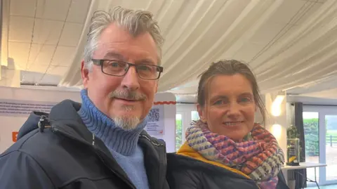 Connor Bennett/BBC A couple smile at the camera in a head and shoulders image of them both, standing side by side. The man, to the left, has blue eyes, grey hair and black framed spectacles, and is wearing a blue polo neck jumper and navy jacket. To the right, the woman has blue eyes, brown hair tied back, a multi-coloured scarf and dark blue jacket.