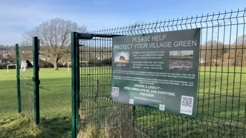 BBC The entrance to Stoke Lodge field. there is a green gate with a large green poster reading 'please help protect your village green'.