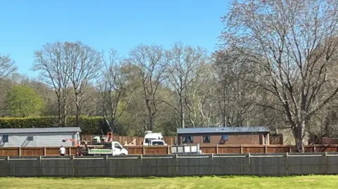 A field with fences in it. In the background, two mobile homes are surrounded by vans and diggers, with people working nearby. There are trees behind this, and a blue sky.