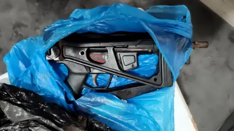 Greater Manchester Police The black MP5 sub-machine gun recovered by police. It is a short, stubby weapon resting side-on in a torn blue plastic carrier bag. The muzzle, which is poking through one side of the bag, looks rusty.