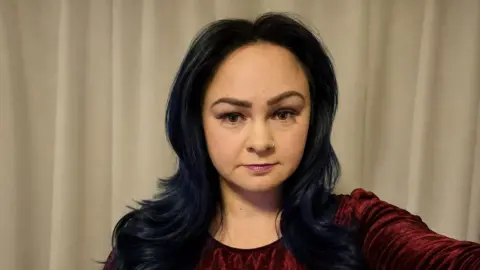 Rebecca Rahore A selfie of a woman with long back hair in a centre parting. She has a small silver hoop nose ring and is wearing a purple velour dress.