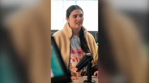 Woman with brown hair stands behind a shop counter with a card machine in view.