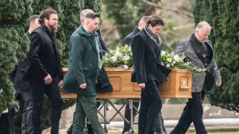 PA Media Seven men with their heads bowed walk alongside a light-brown wooden coffin, which is supported by a metal trolley, against a backdrop of a church-yard.