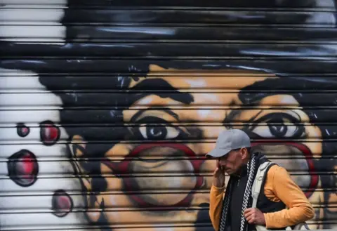 Amr Abdallah Dalsh / Reuters An Egyptian man walks past a shop in the morning. On the shutters is painted a mural of a woman wearing red glasses - Saturday 1 November 2025.
