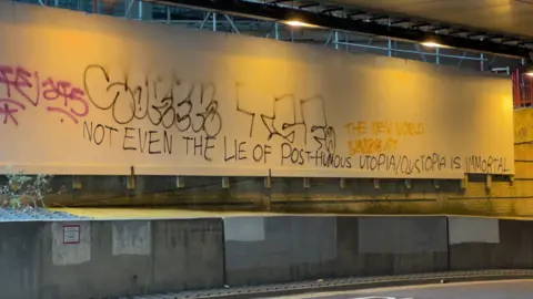 The wall beneath a bridge is covered with layered graffiti and bold handwritten text. The tunnel lighting casts a warm yellow glow across the scene. Metal beams and construction elements frame the space above the graffiti.